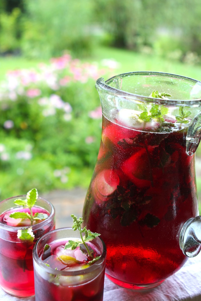 A pitcher of Hibiscus Mint Herbal Iced Tea on a table next to two full glasses garnished with sprigs of mint.