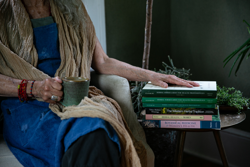 A person holding a mug sits next to a stack of some of the best herbal medicine books for herbalists.