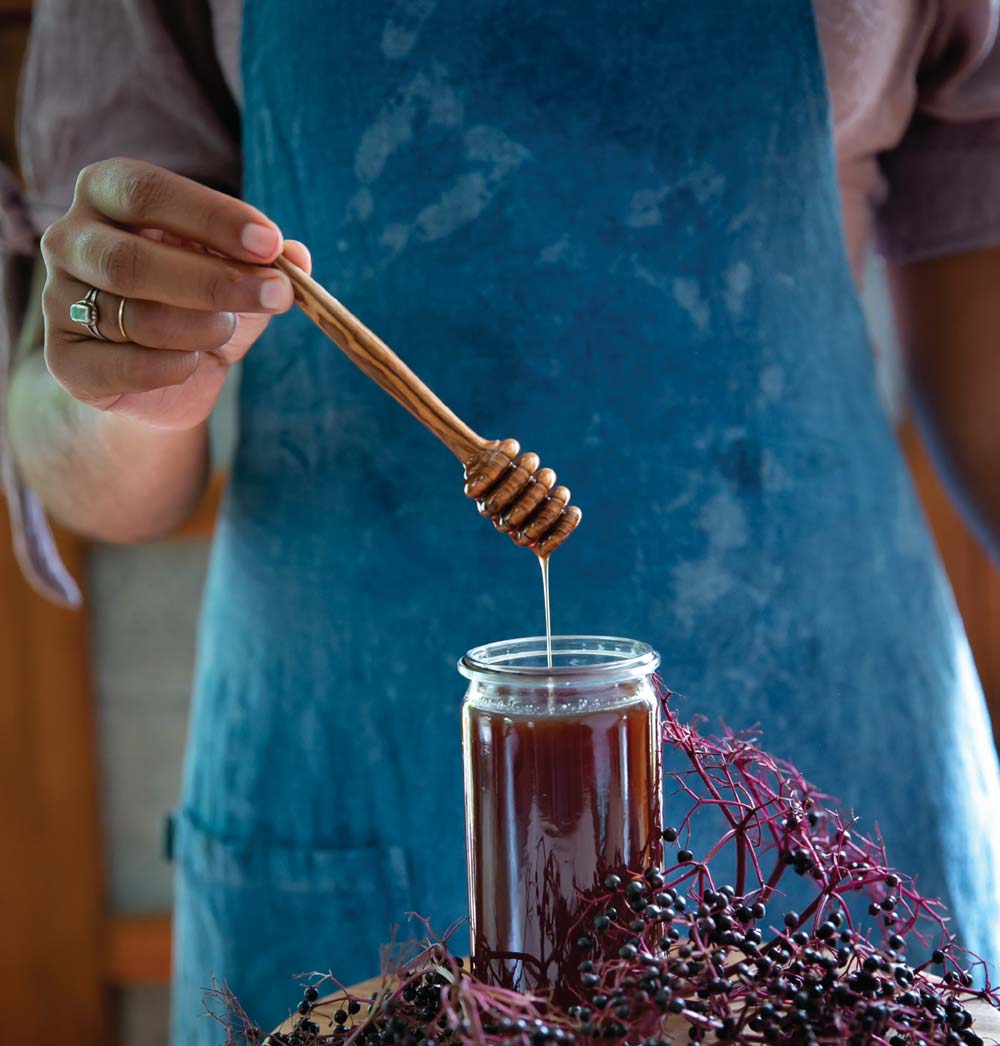 A person holds a honey dropper over a jar of elderberry infused honey.